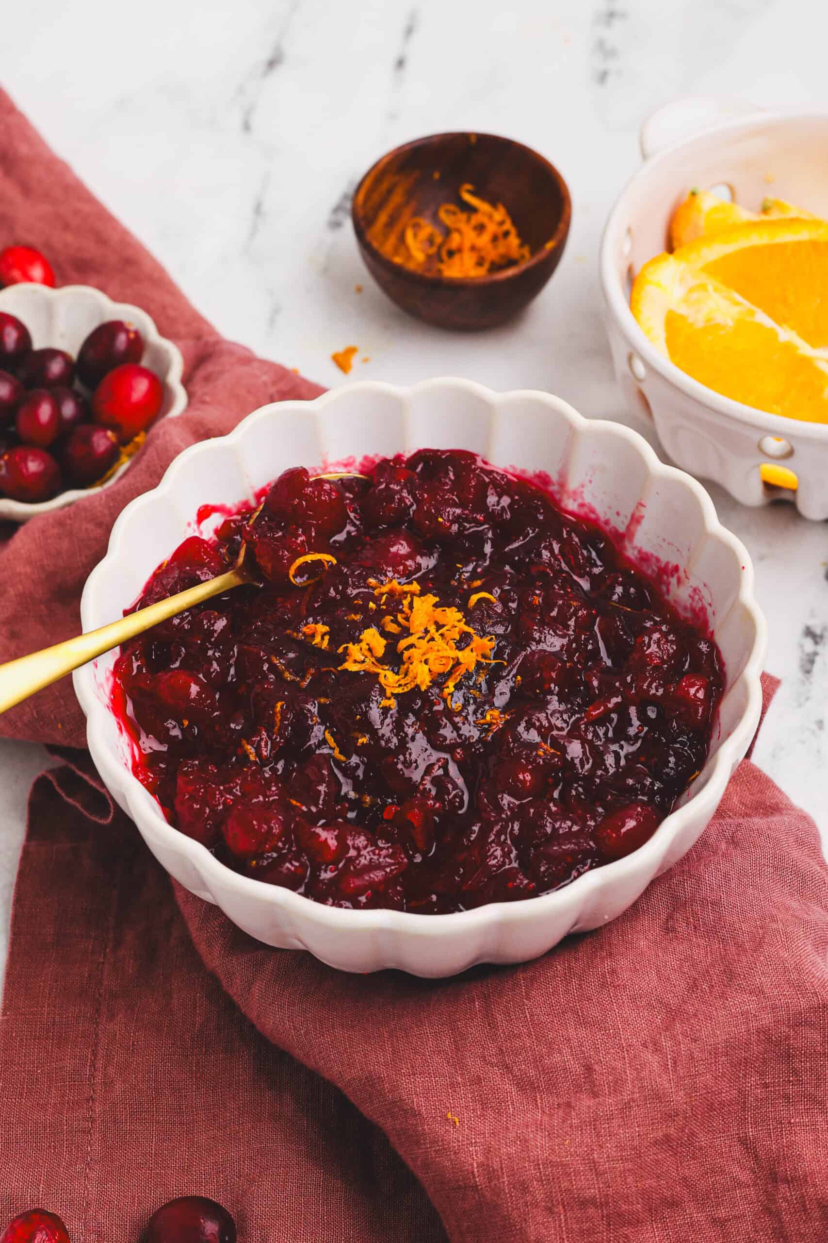 Homemade cranberry orange sauce served in a white bowl with fresh cranberries and orange slices on a rustic cloth.