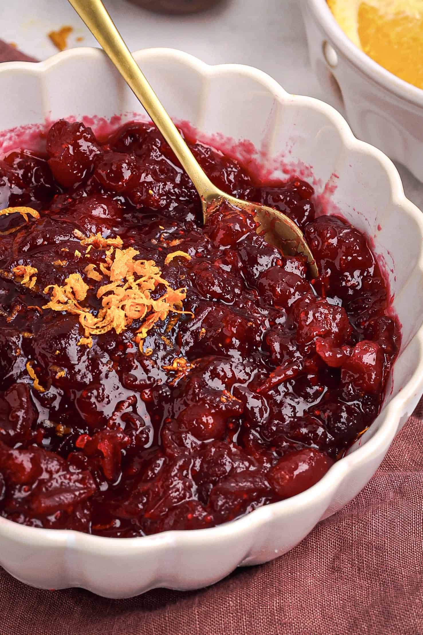 Close-up of homemade cranberry orange sauce in a white bowl with a gold spoon and fresh orange zest on top.