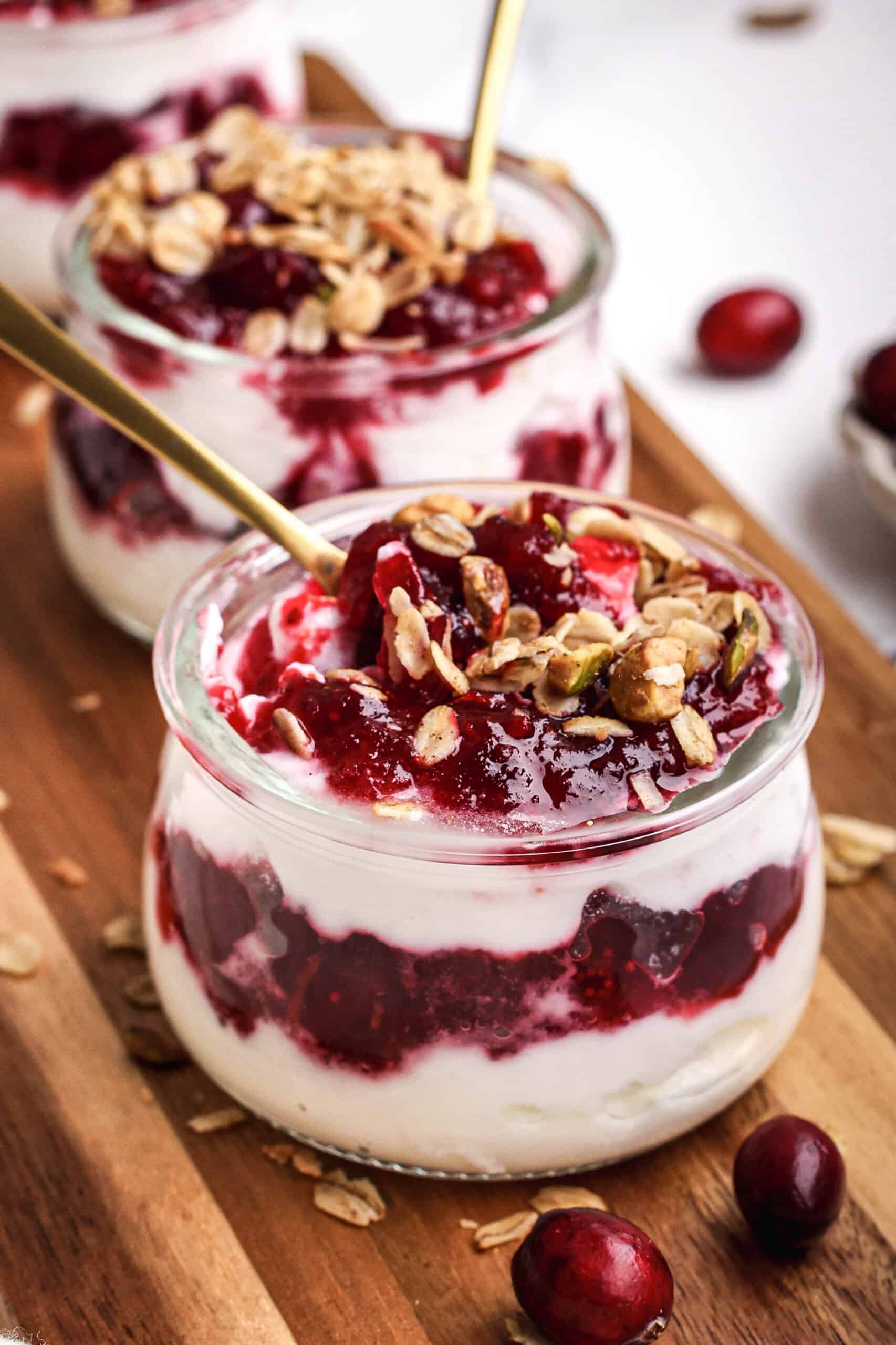 Cranberry sauce granola parfait layered with creamy yogurt, tart cranberry sauce, and crunchy granola in a glass jar, topped with oats and served on a wooden board with fresh cranberries.