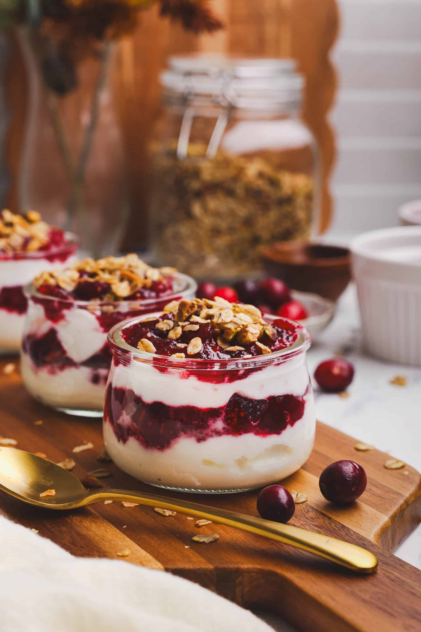 Cranberry granola parfaits layered with creamy yogurt, cranberry sauce, and crunchy granola in glass jars, served on a wooden board with fresh cranberries and a gold spoon.