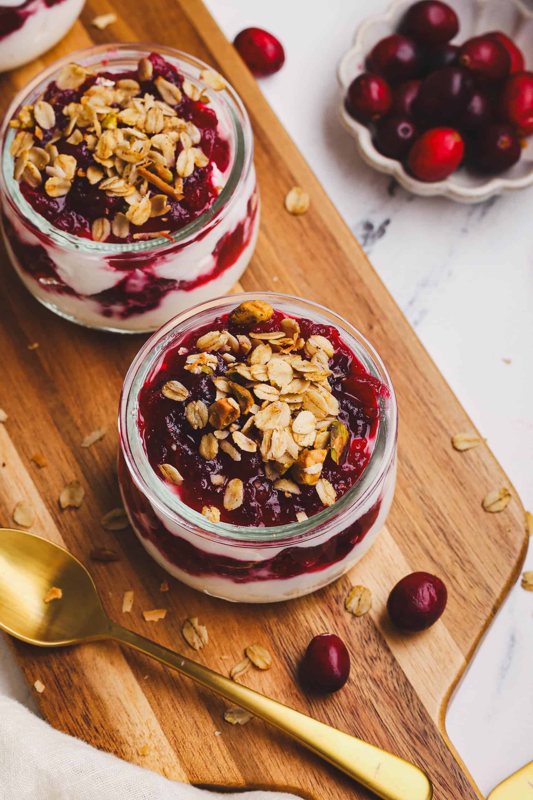 Overhead view of cranberry granola parfaits layered with yogurt and cranberry sauce, topped with crunchy granola, served on a wooden board with fresh cranberries and a gold spoon.