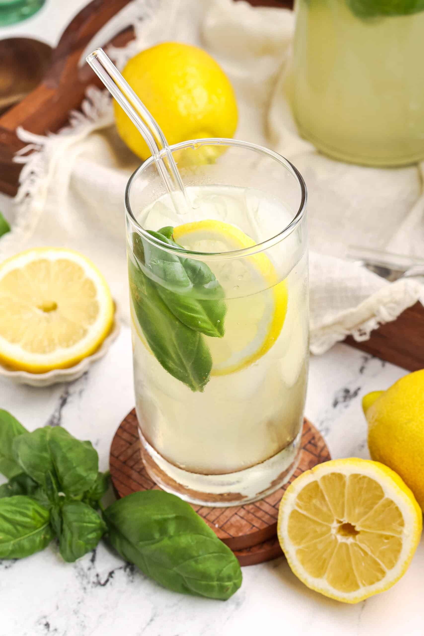 Glass of fresh basil lemonade with lemon slice and basil, styled with whole lemons and herbs on a marble surface.
