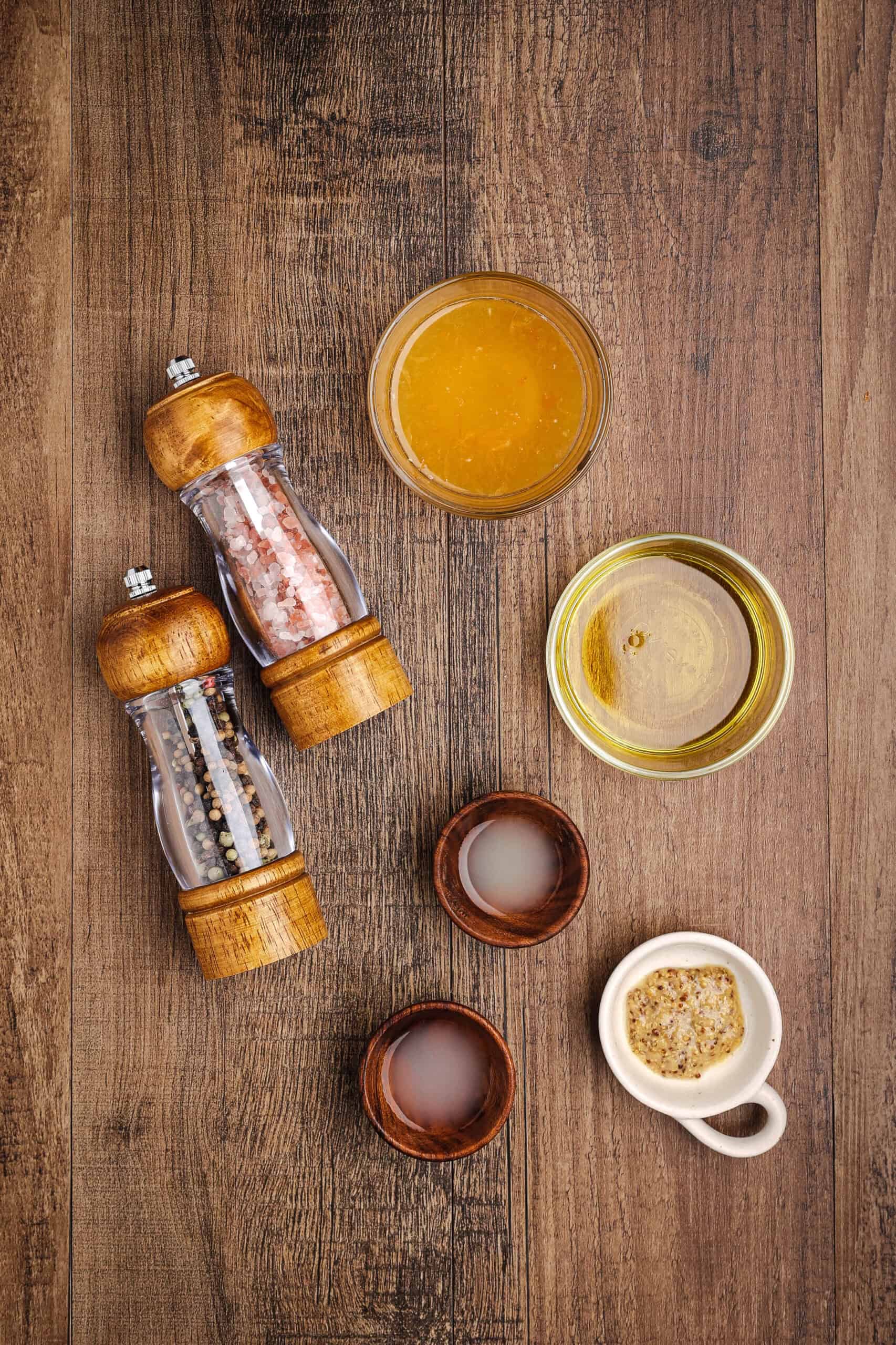Flat lay of citrus dressing ingredients including olive oil, orange marmalade, Dijon mustard, lemon juice, lime juice, salt, and pepper on a rustic wooden surface.