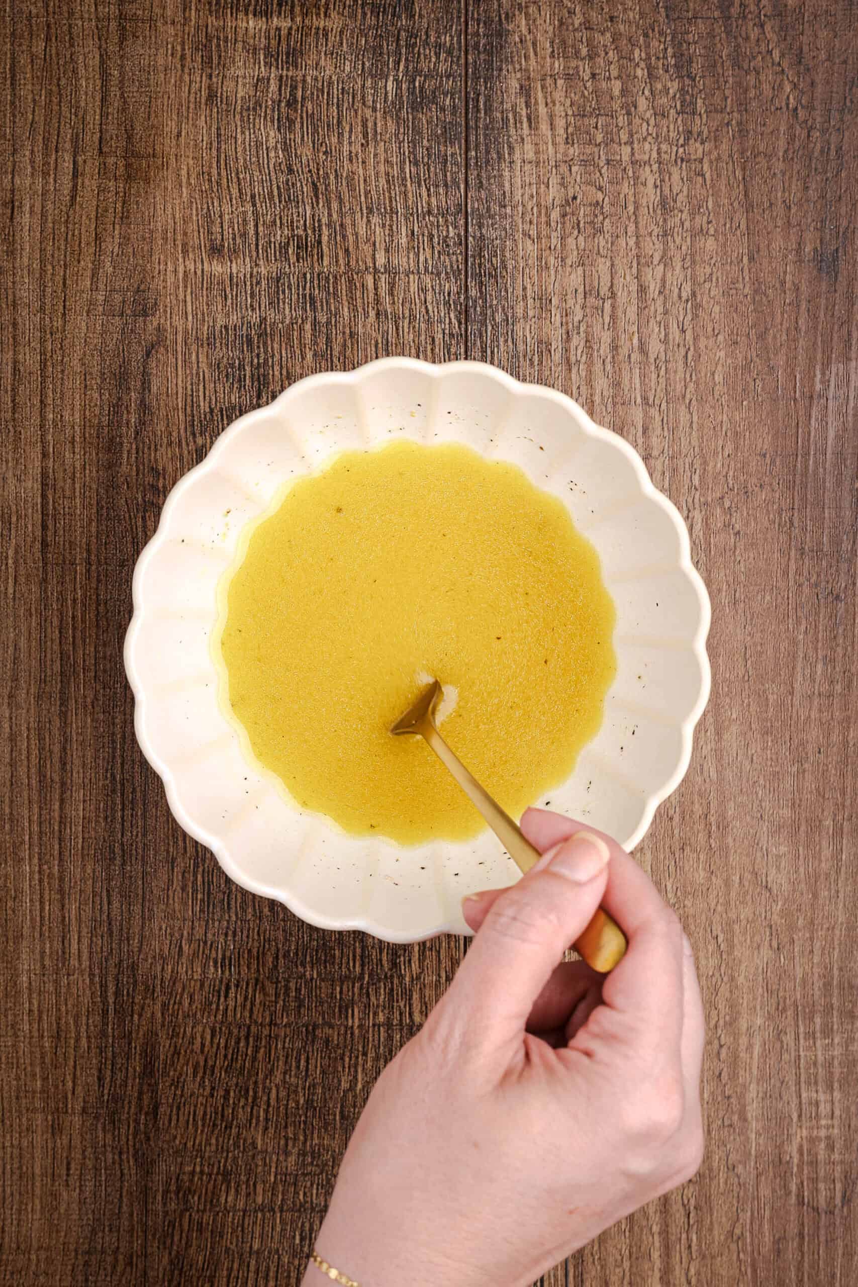Hand whisking homemade citrus salad dressing in a scalloped ceramic bowl with a gold spoon on a wooden table.