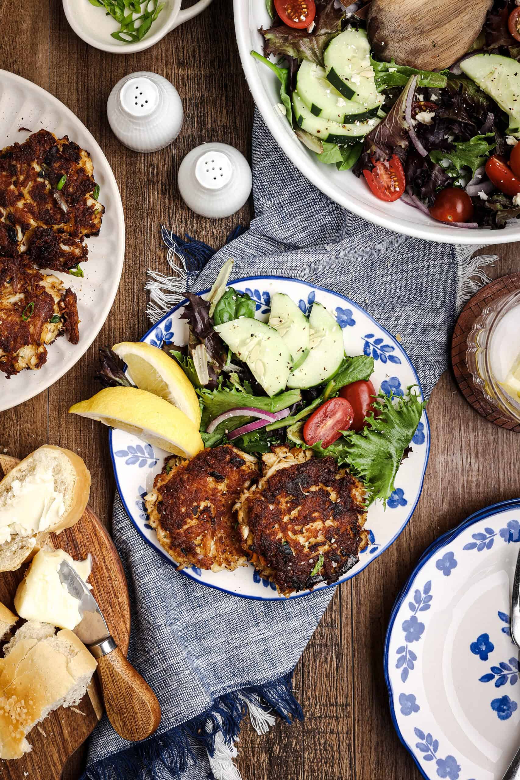 Overhead hero shot of homemade crab cake salad with crispy crab cakes, mixed greens, cucumbers, tomatoes, lemon wedges, rustic bread, and coastal-inspired blue floral plates on a wooden table.