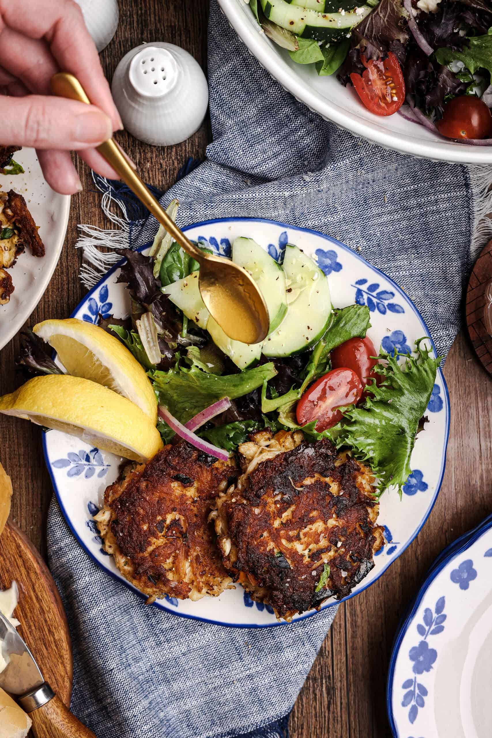 Overhead photo of crispy homemade crab cakes served with mixed greens, cucumbers, tomatoes, red onion, and lemon wedges while citrus dressing is drizzled over the salad with a gold spoon.