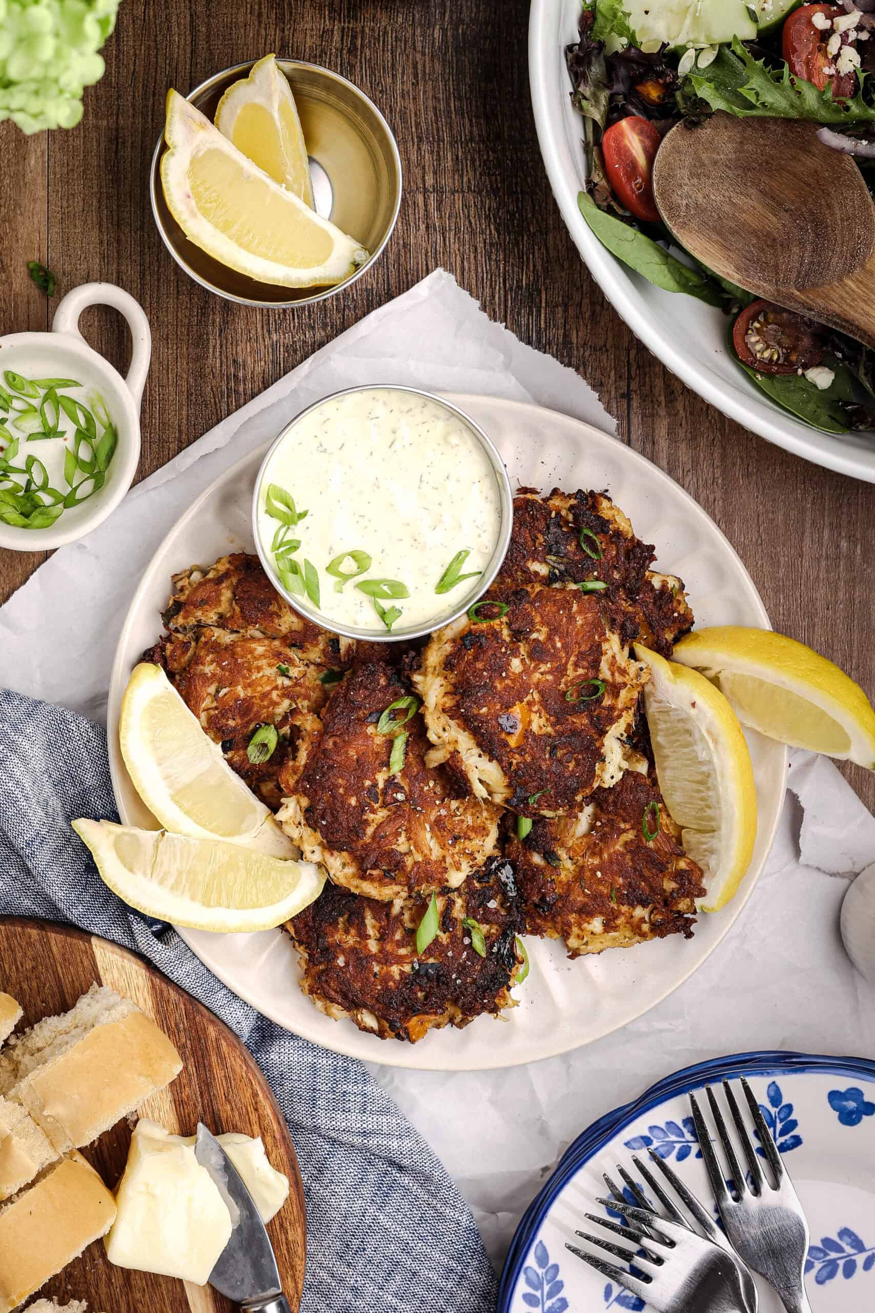Overhead hero shot of crispy homemade crab cakes served with tartar sauce, lemon wedges, fresh salad, and rustic bread on a wooden table.