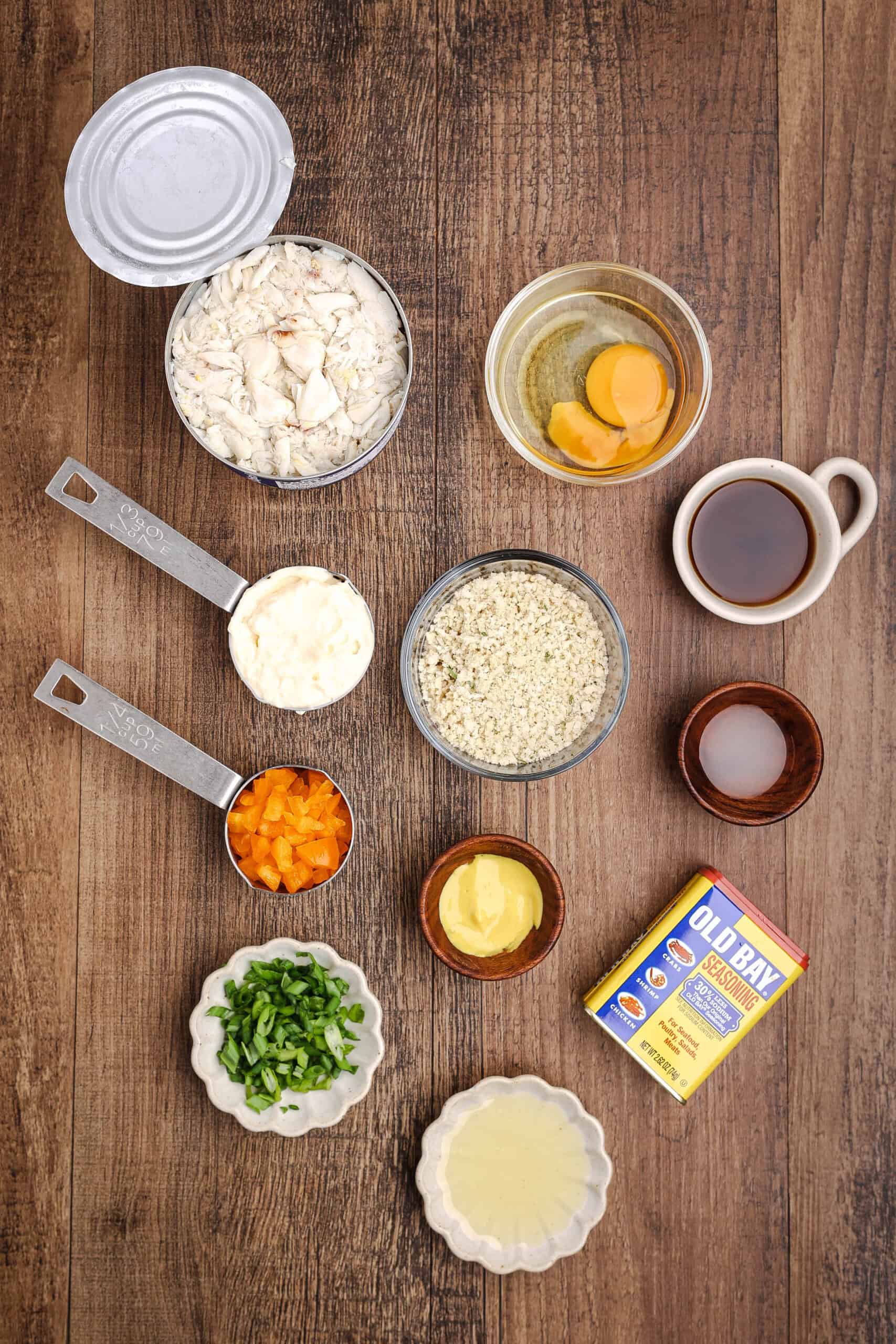 Flat lay of ingredients for homemade crab cakes including lump crab meat, egg, mayonnaise, breadcrumbs, Worcestershire sauce, Dijon mustard, green onions, diced bell pepper, lemon juice, and Old Bay seasoning on a wooden surface.