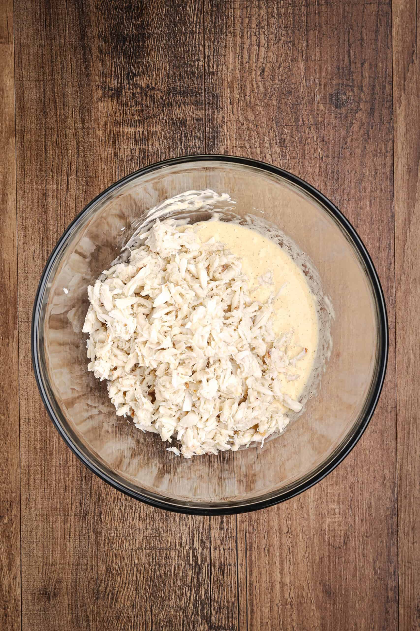Lump crab meat being added to a creamy crab cake mixture in a large glass mixing bowl during recipe preparation.