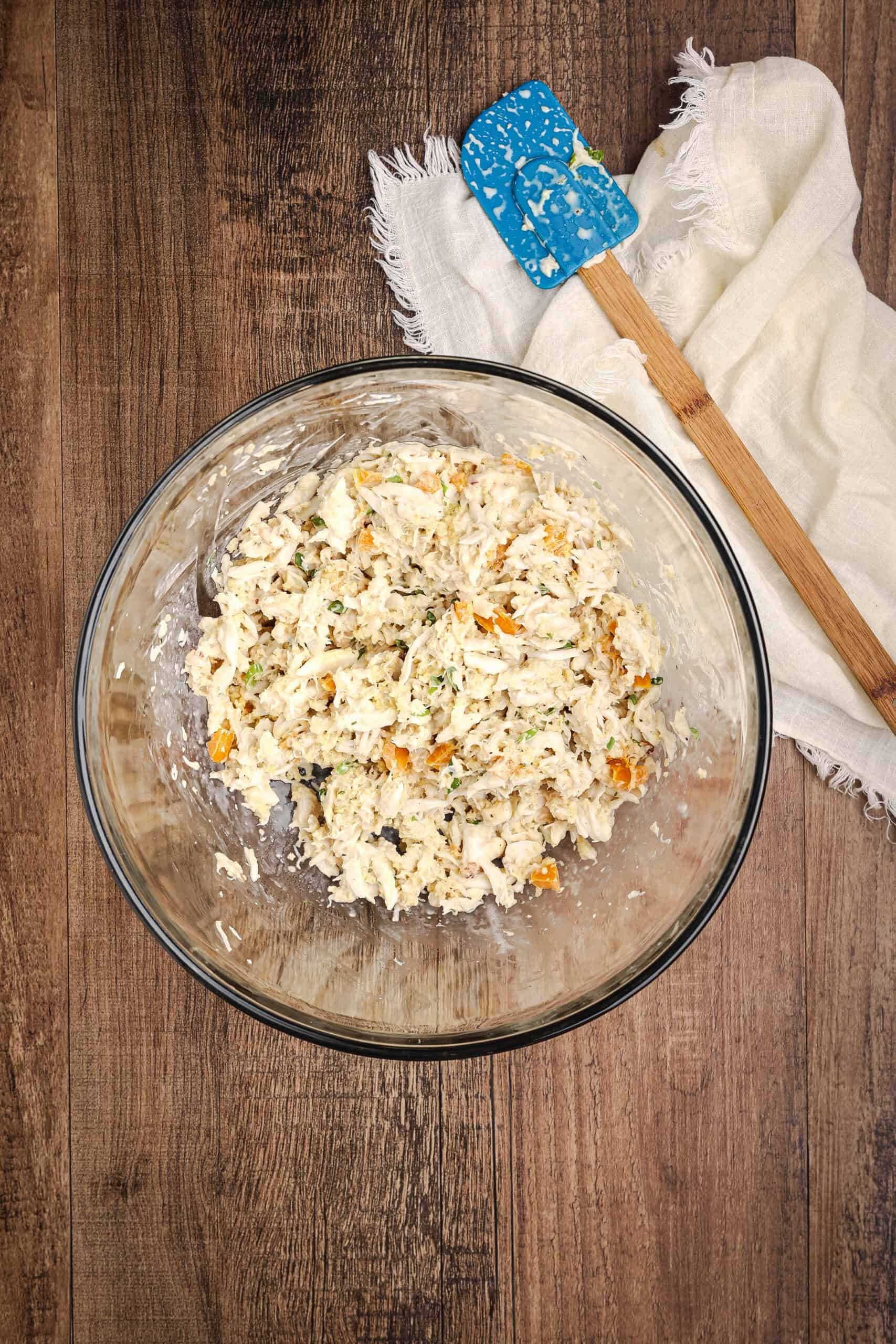 Homemade crab cake mixture combined in a glass bowl with diced bell peppers and green onions beside a blue spatula on a rustic wooden table.