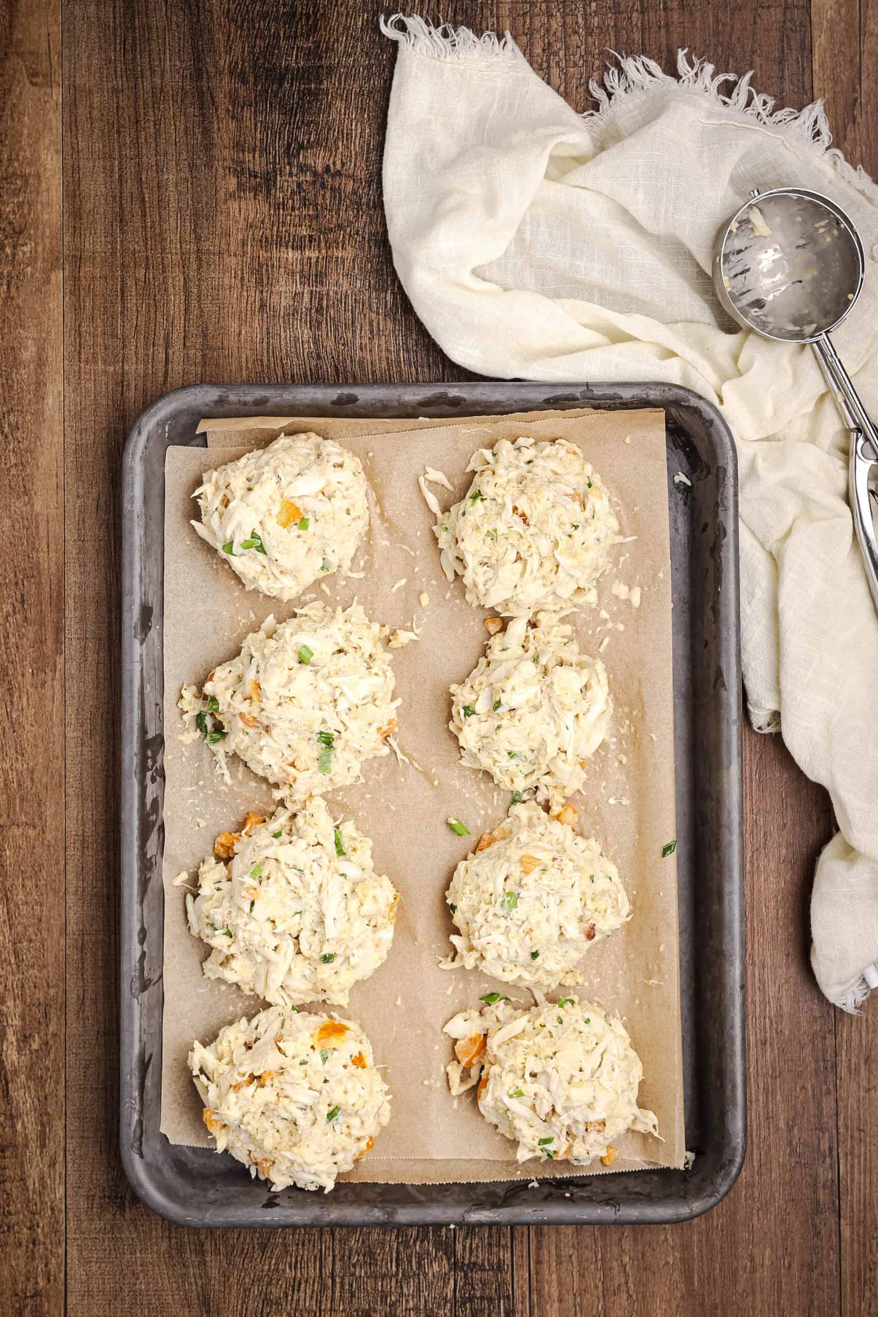 Uncooked crab cakes shaped and arranged on a parchment-lined baking tray before cooking, styled with a linen cloth and scoop.