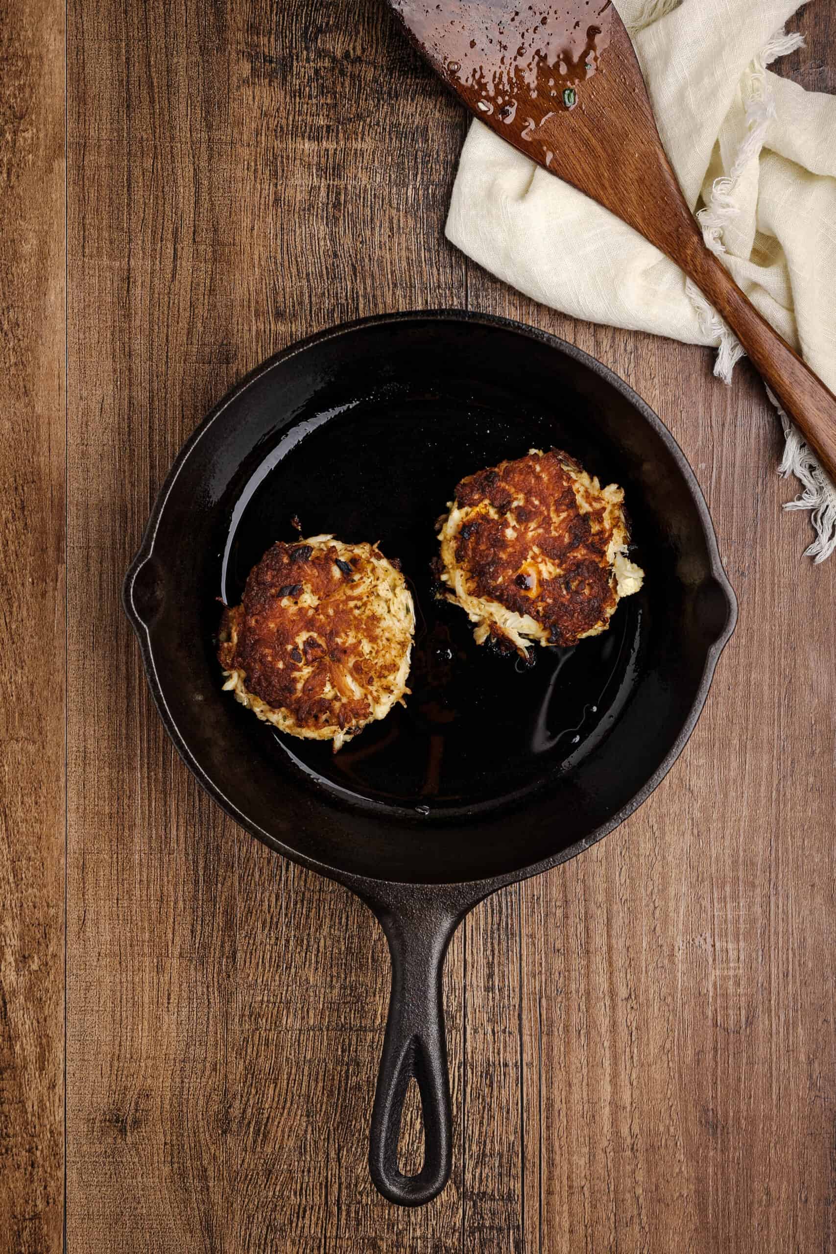 Golden brown crab cakes cooking in a cast iron skillet with crispy seared edges on a rustic wooden background.