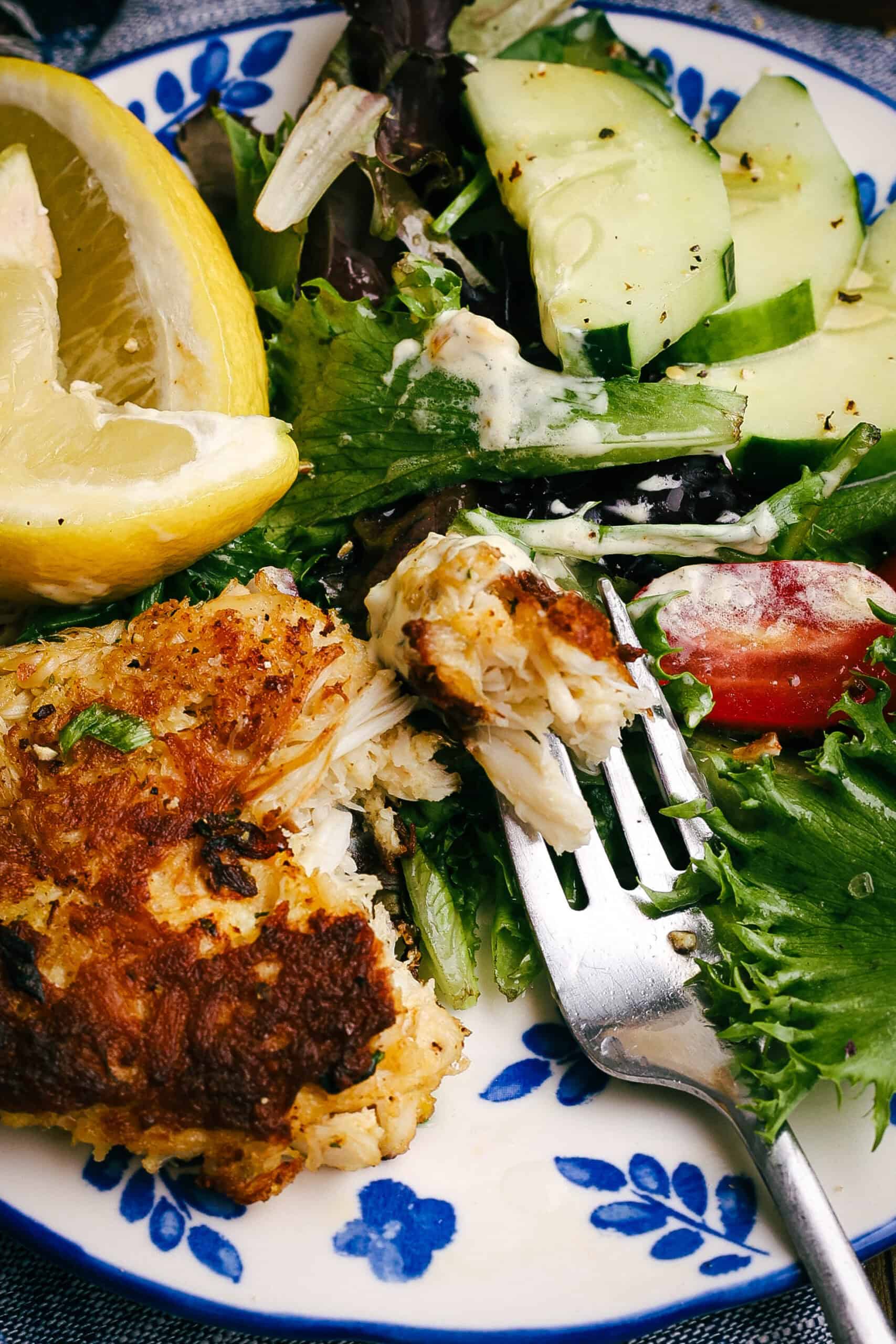 Close-up of flaky homemade crab cake served with fresh salad greens, cucumber slices, cherry tomatoes, creamy dressing, and a lemon wedge on a blue floral plate.