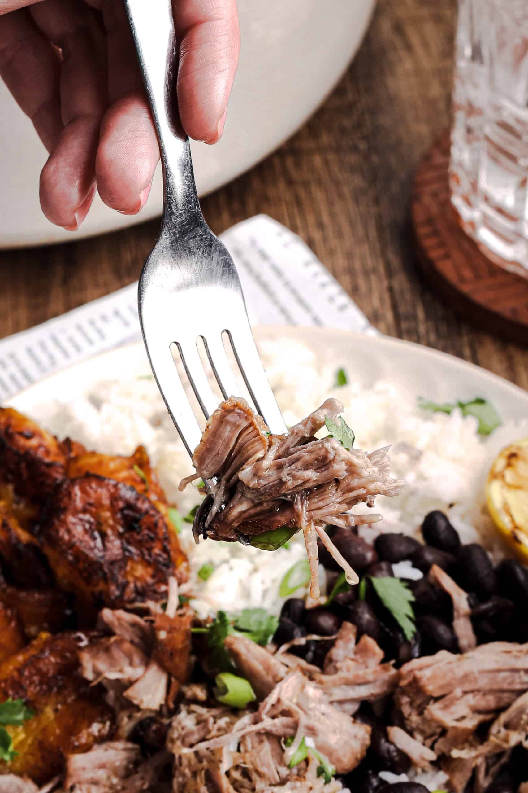 Fork holding a bite of juicy Cuban mojo pork roast above a plate of rice, black beans, and fried sweet plantains.