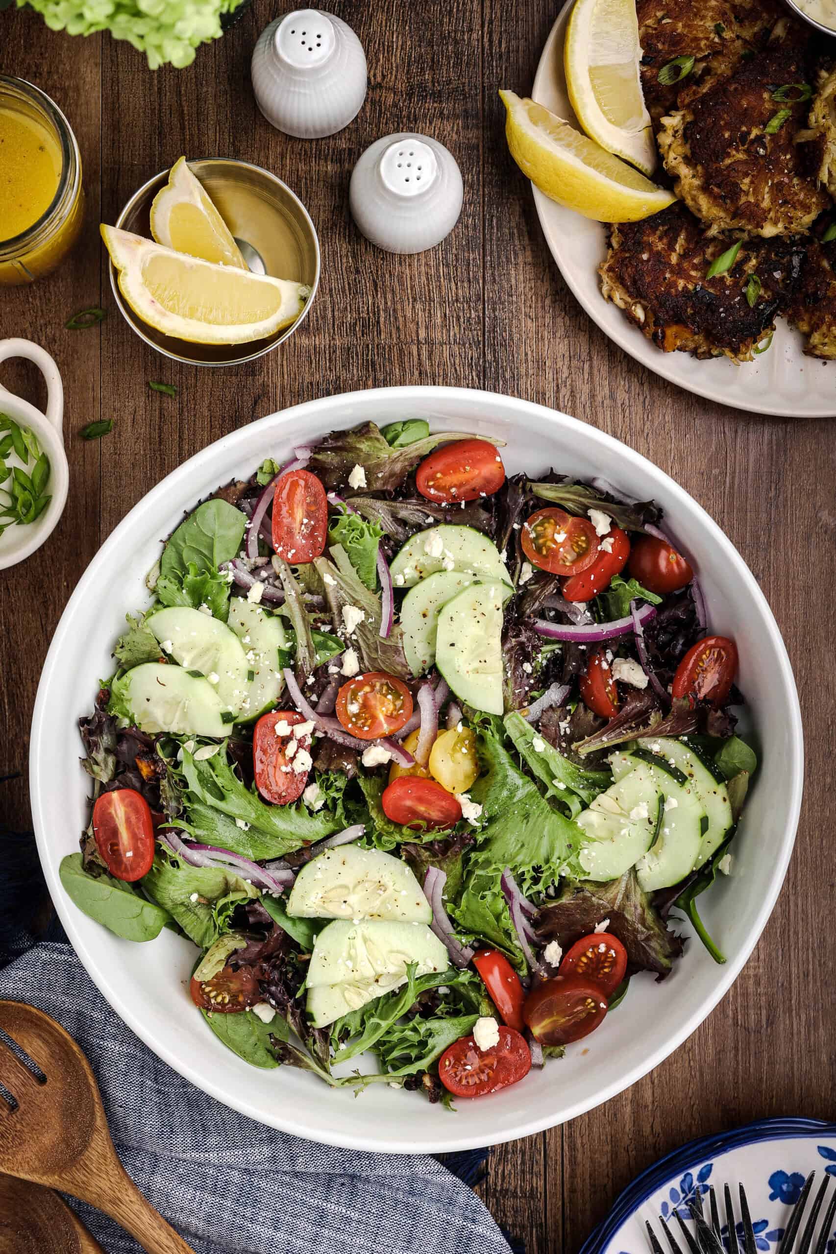 Large bowl of fresh mixed green salad with cucumbers, cherry tomatoes, red onion, feta cheese, and citrus dressing served alongside crab cakes and lemon wedges.