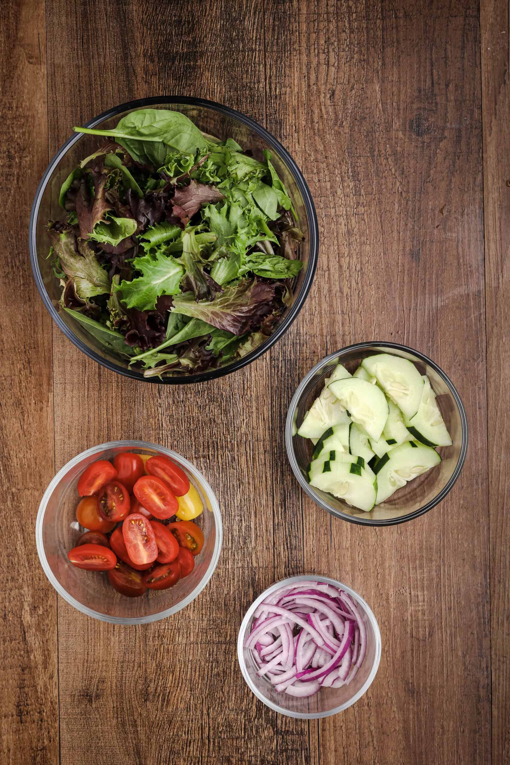 Overhead view of salad ingredients including mixed greens, sliced cucumbers, cherry tomatoes, and red onion arranged in glass bowls on a wooden background.