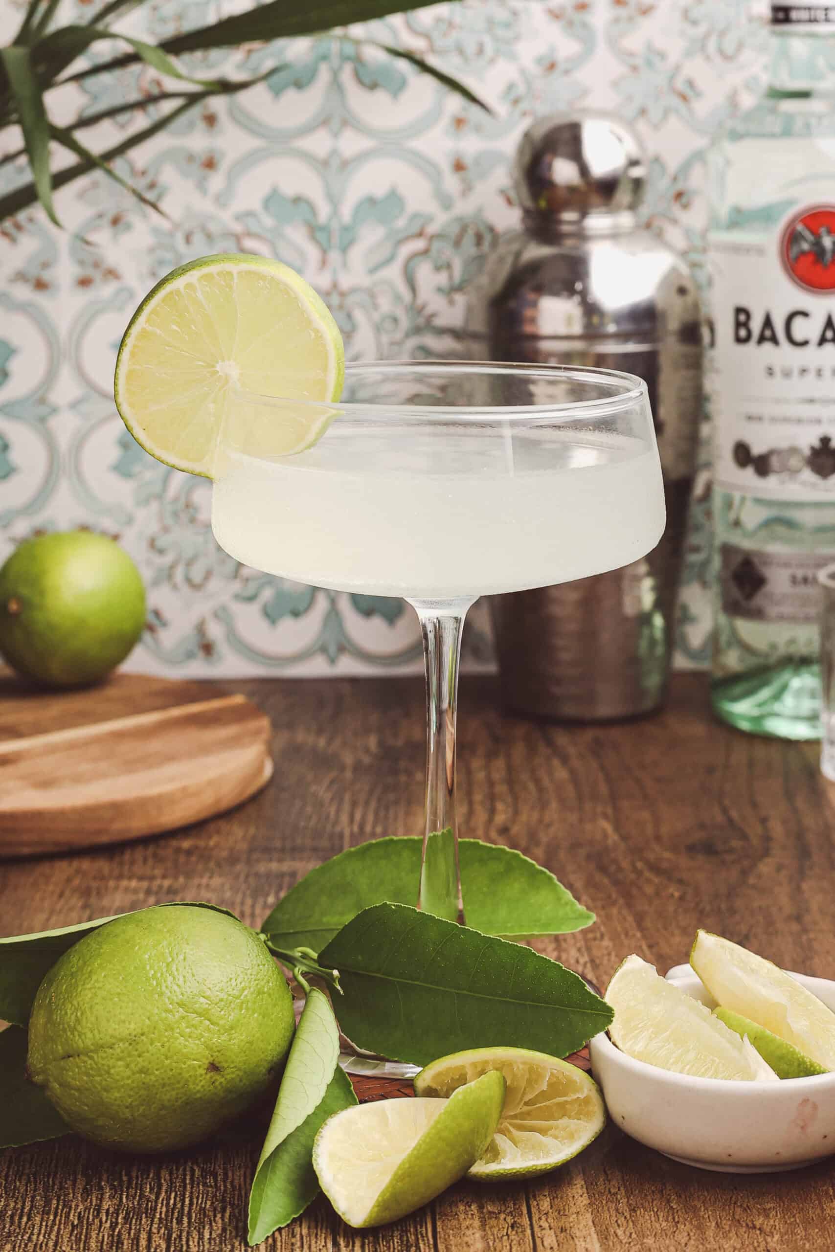 Classic daiquiri cocktail garnished with a lime wheel in a coupe glass, styled with fresh limes, citrus leaves, and Bacardí rum on a wooden table.