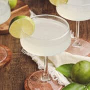 Three classic daiquiris in coupe glasses arranged on a wooden table with fresh Florida limes and linen napkins.