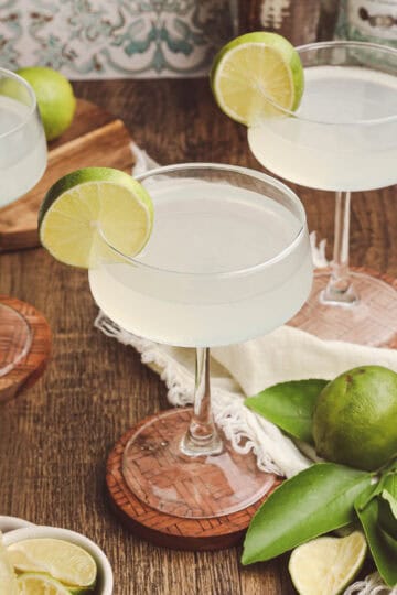 Three classic daiquiris in coupe glasses arranged on a wooden table with fresh Florida limes and linen napkins.