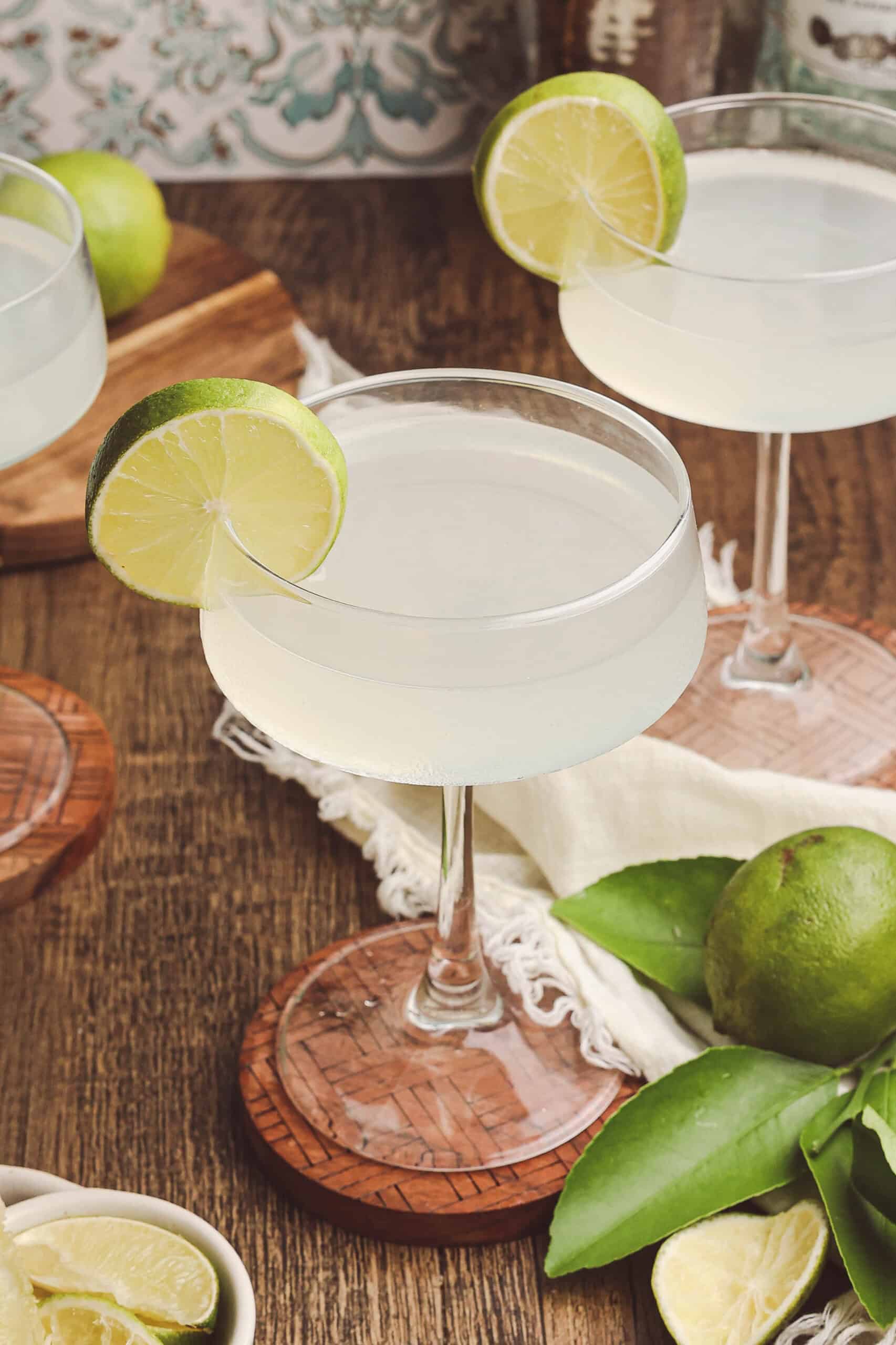 Three classic daiquiris in coupe glasses arranged on a wooden table with fresh Florida limes and linen napkins.