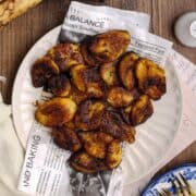 Overhead view of fried sweet plantains served on a white plate lined with newspaper-style parchment, surrounded by ripe plantains, a cast iron skillet, and a linen cloth on a rustic wooden table.