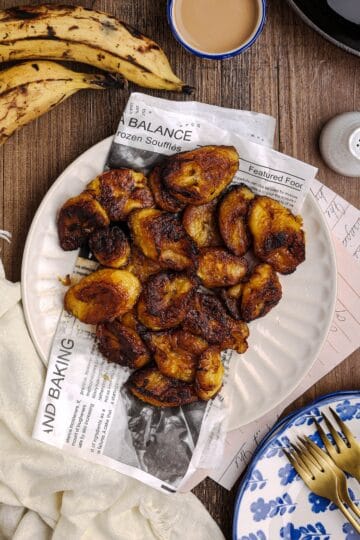 Overhead view of fried sweet plantains served on a white plate lined with newspaper-style parchment, surrounded by ripe plantains, a cast iron skillet, and a linen cloth on a rustic wooden table.