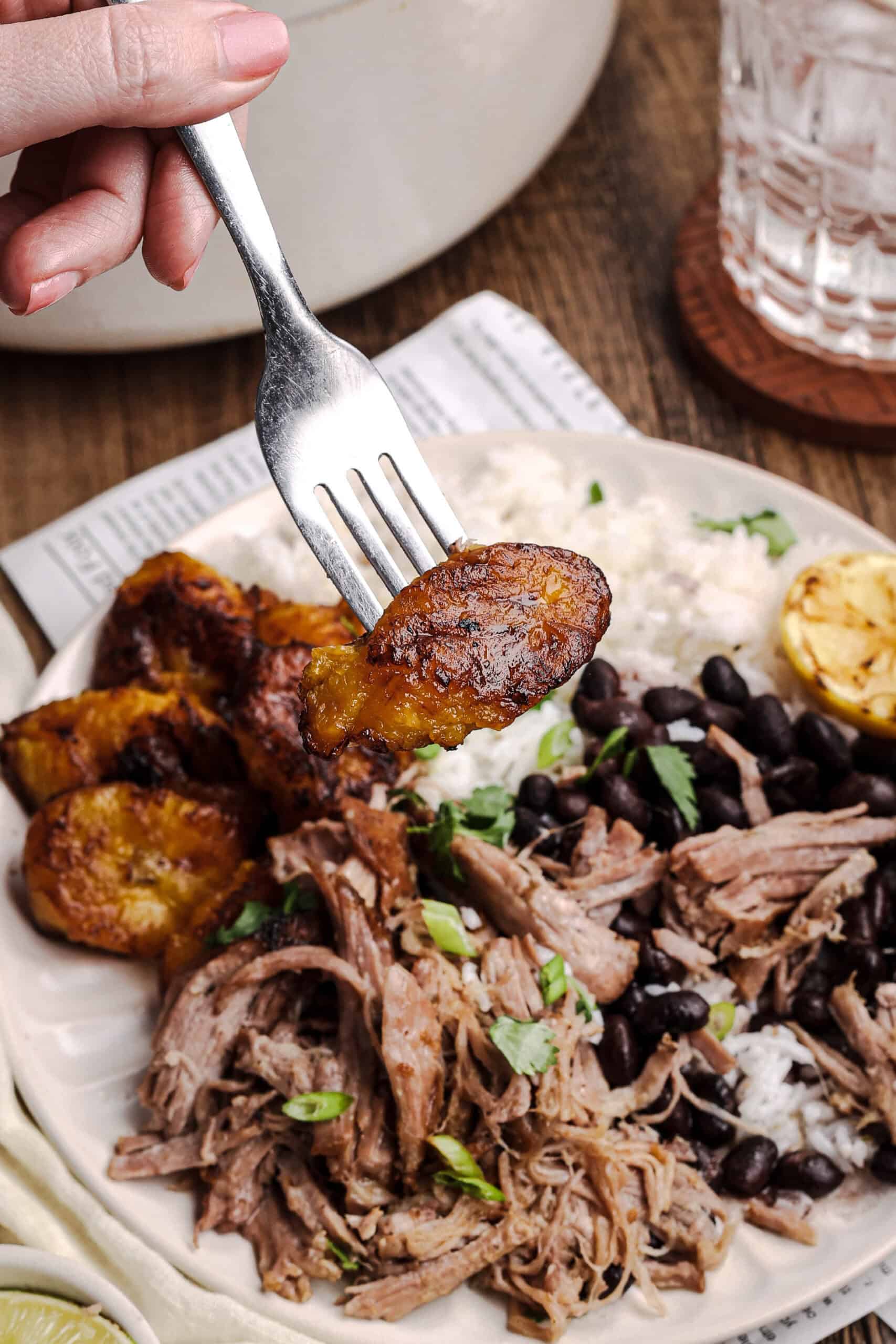 Fork holding a caramelized fried plantain above a plate of mojo pork, black beans, and rice, showcasing a classic Cuban-style meal.