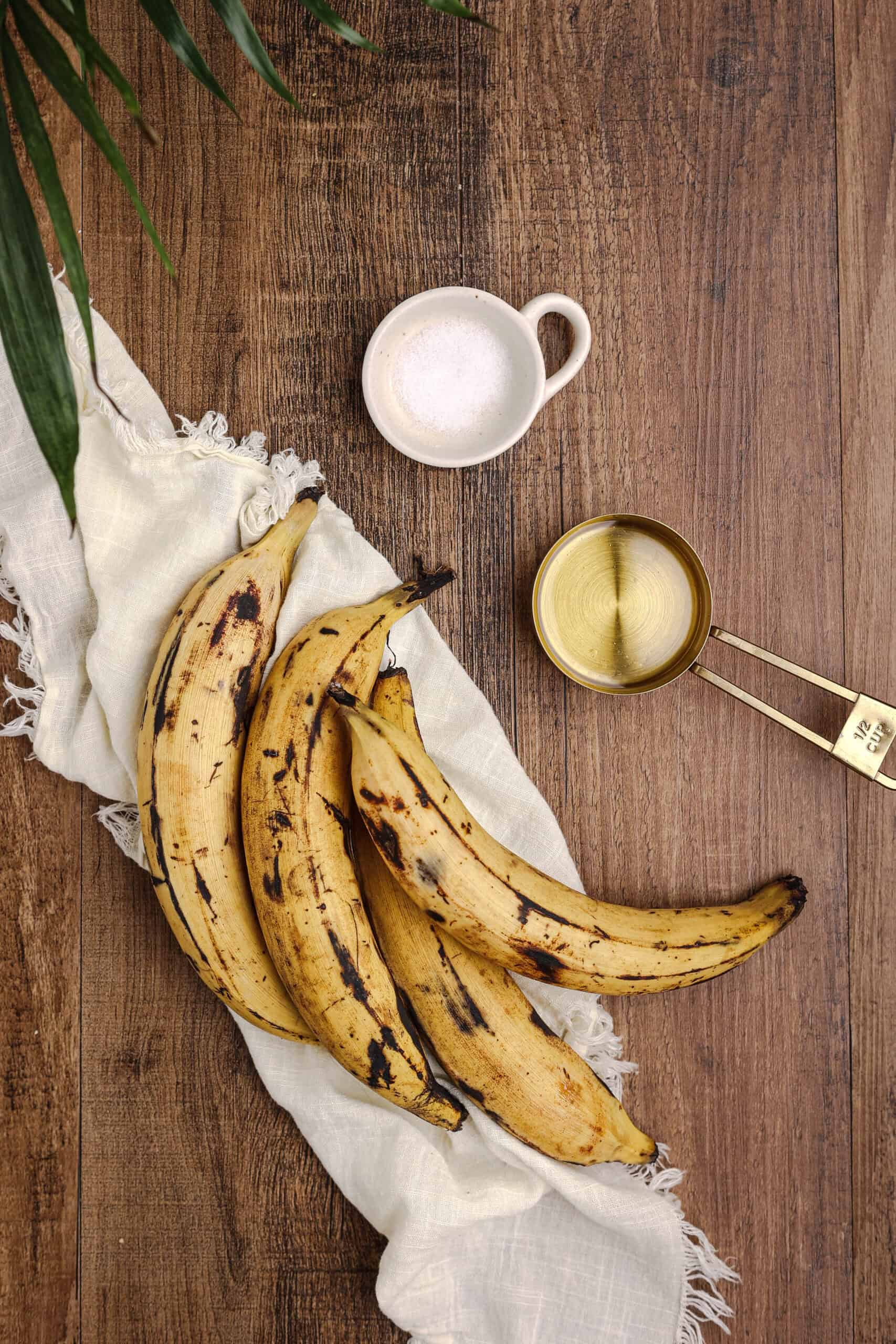 Ripe plantains, oil, and salt arranged on a rustic wooden table with linen cloth, showing the simple ingredients needed for fried sweet plantains.