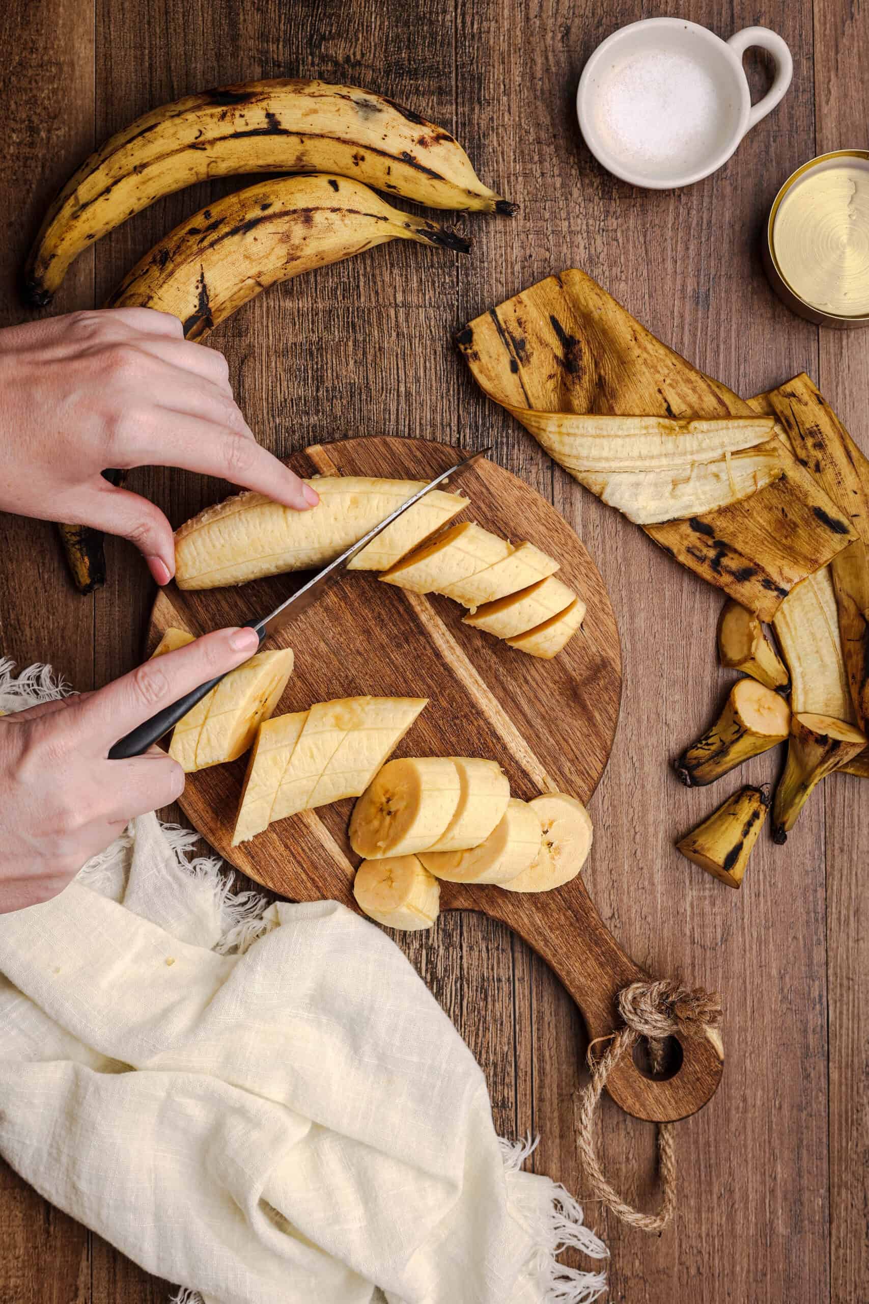 Hands slicing ripe plantains into diagonal pieces on a wooden cutting board for making fried sweet plantains.