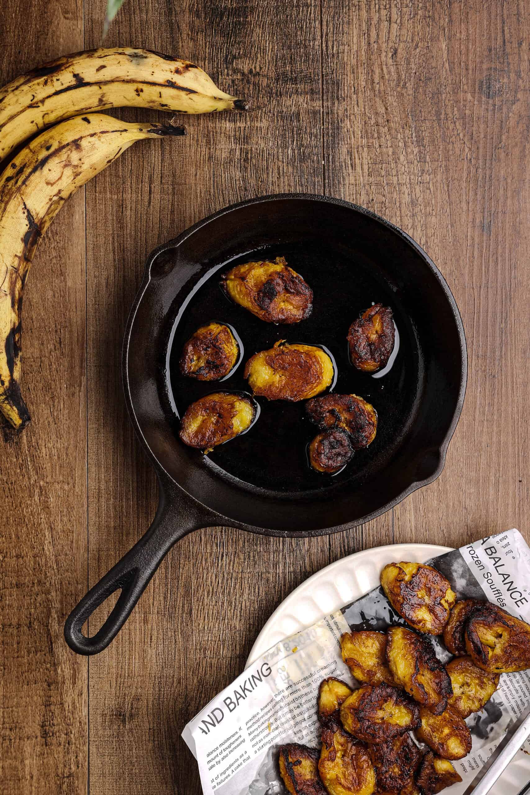 Sweet plantain slices frying in a cast iron skillet with hot oil, developing their signature golden caramelized exterior.
