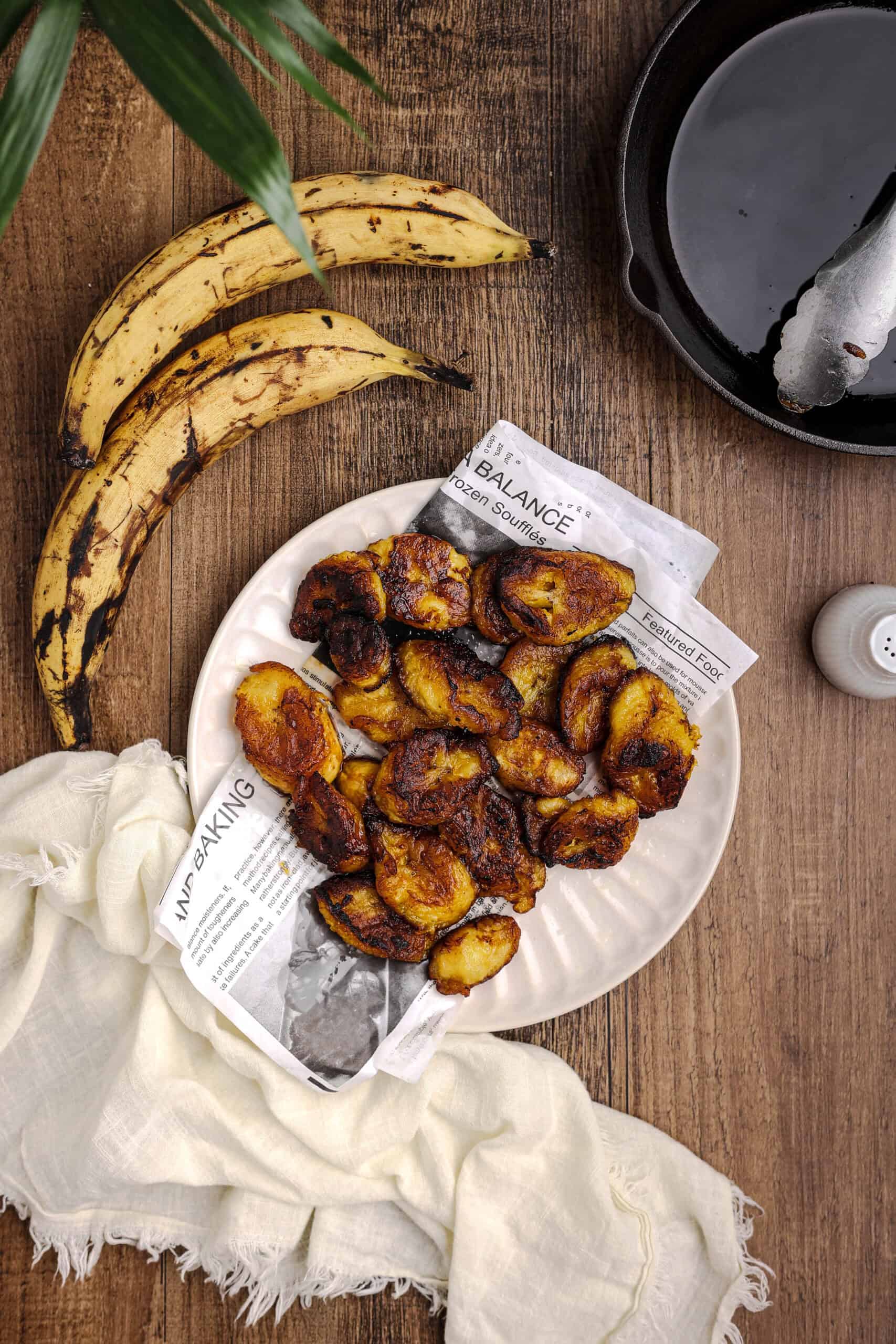 Overhead view of fried sweet plantains served on a white plate lined with newspaper-style parchment, surrounded by ripe plantains, a cast iron skillet, and a linen cloth on a rustic wooden table.