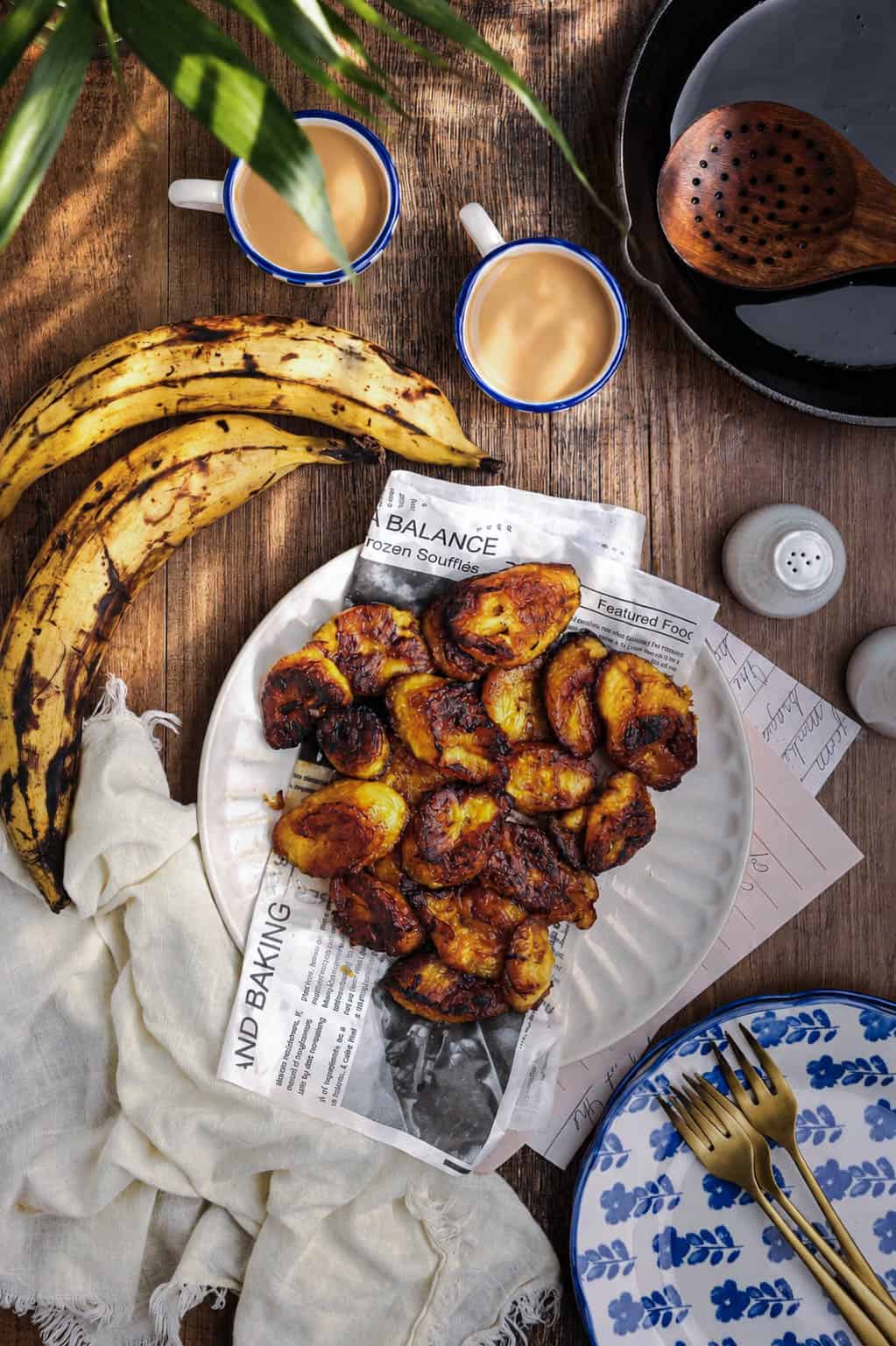 Caramelized fried plantains on a white serving plate with café con leche, ripe plantains, and vintage-inspired table styling on a wooden tabletop.